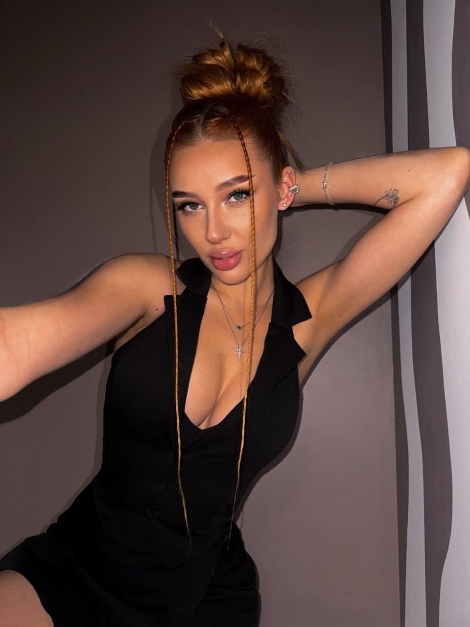 Against a striped wall, she poses with an arm raised, wearing a black plunging neckline top and cross necklace.