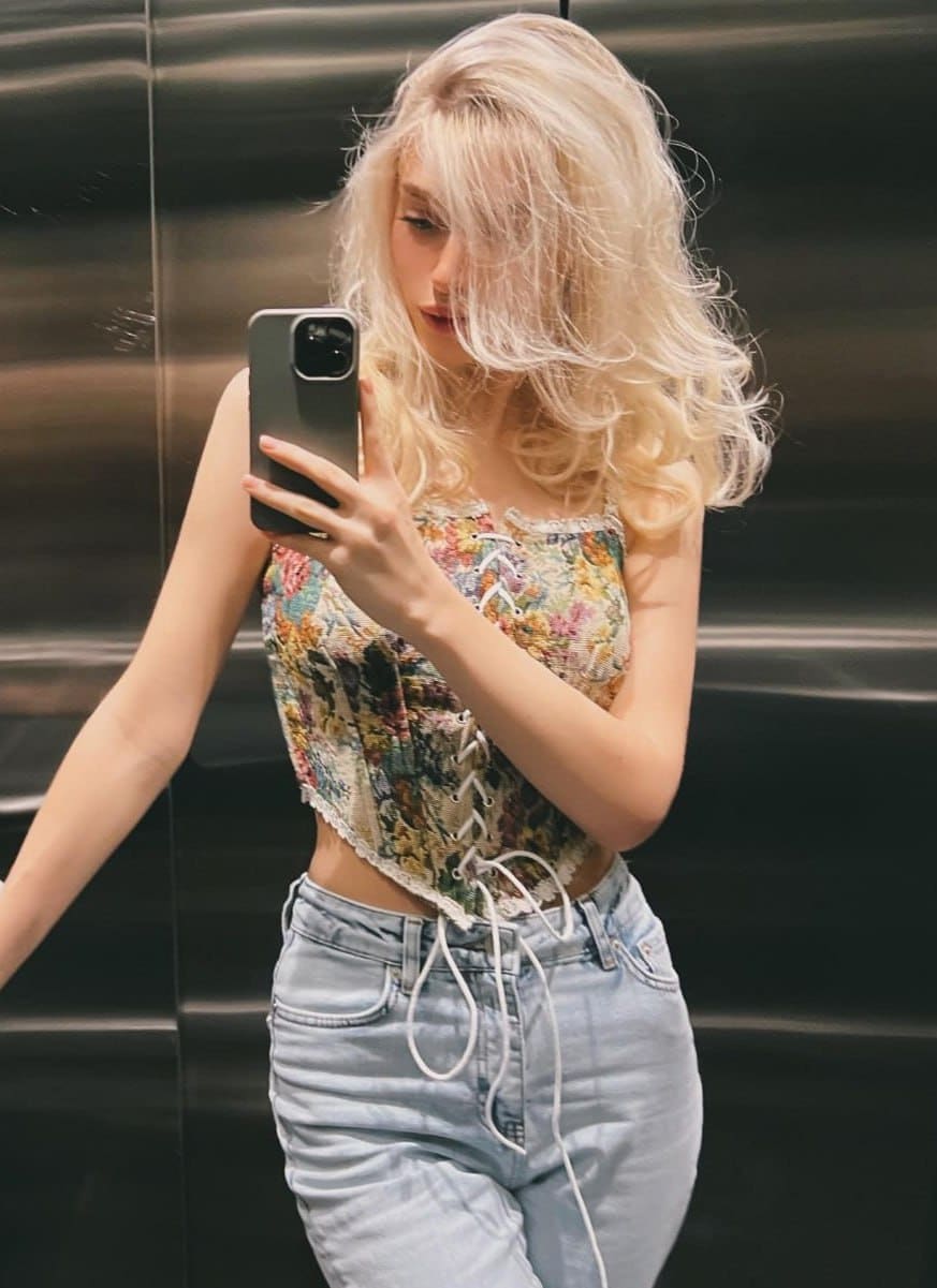 In an elevator mirror selfie, Laura wears a floral lace-up corset top and light-wash blue jeans.