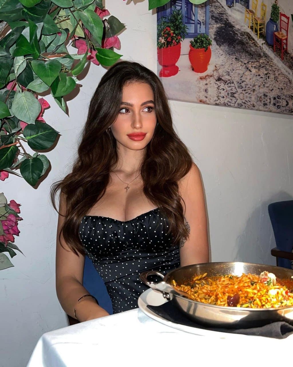 Sophia sits at a table with a pan of food, wearing a black polka-dot strapless dress, against a white wall decorated with greenery and a painting.
