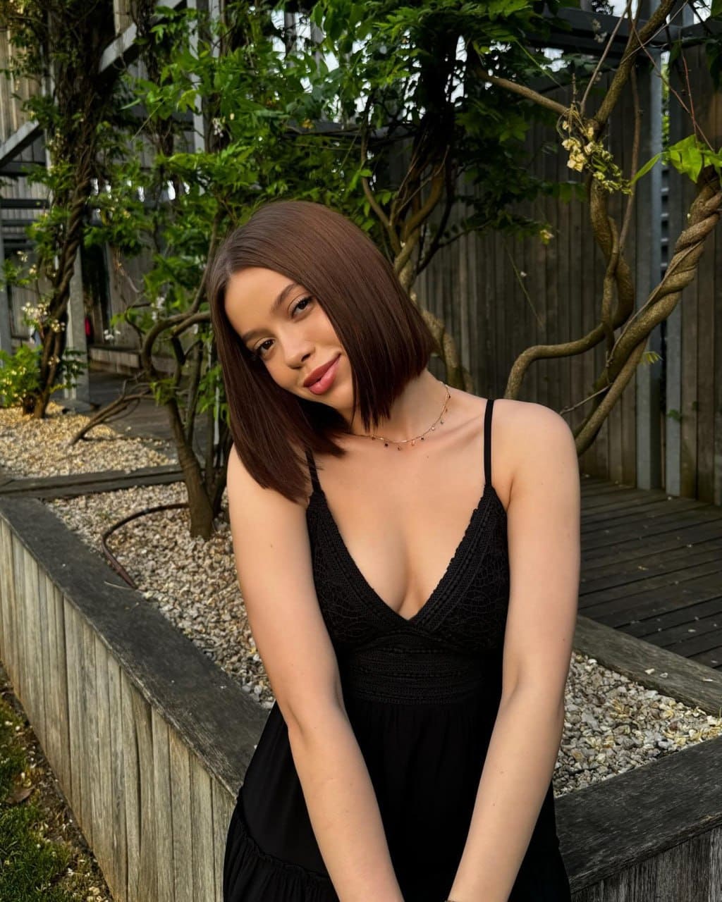 Juliana tilts her head in a black spaghetti-strap dress with a deep V-neck, standing outdoors by a wooden planter and climbing vines.