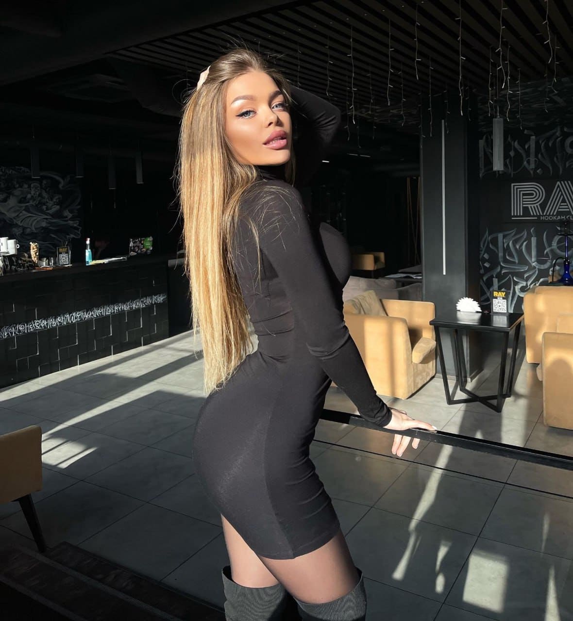 In a lounge setting, Halia stands in profile wearing a tight black mini dress and thigh-high boots, one hand resting on her head.