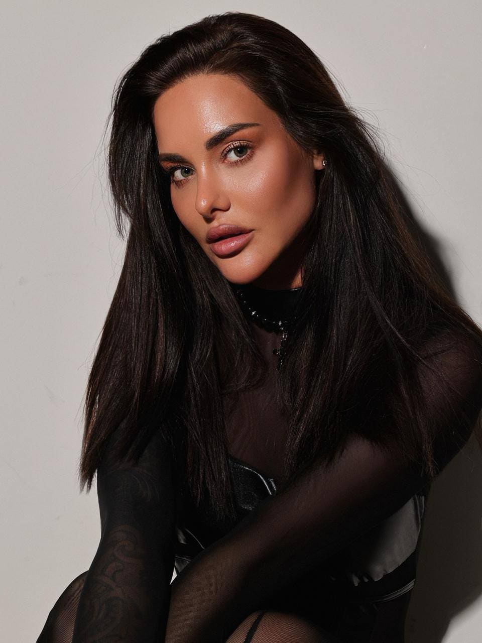 Eliana poses with arms crossed against a plain background, wearing a sheer black mesh top over a dark undergarment.