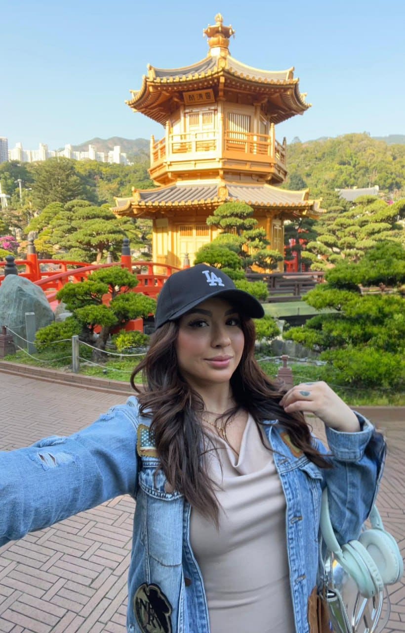 Rosa takes an outdoor selfie in a black cap and denim jacket, holding mint headphones, with a golden pagoda and red bridge behind her.