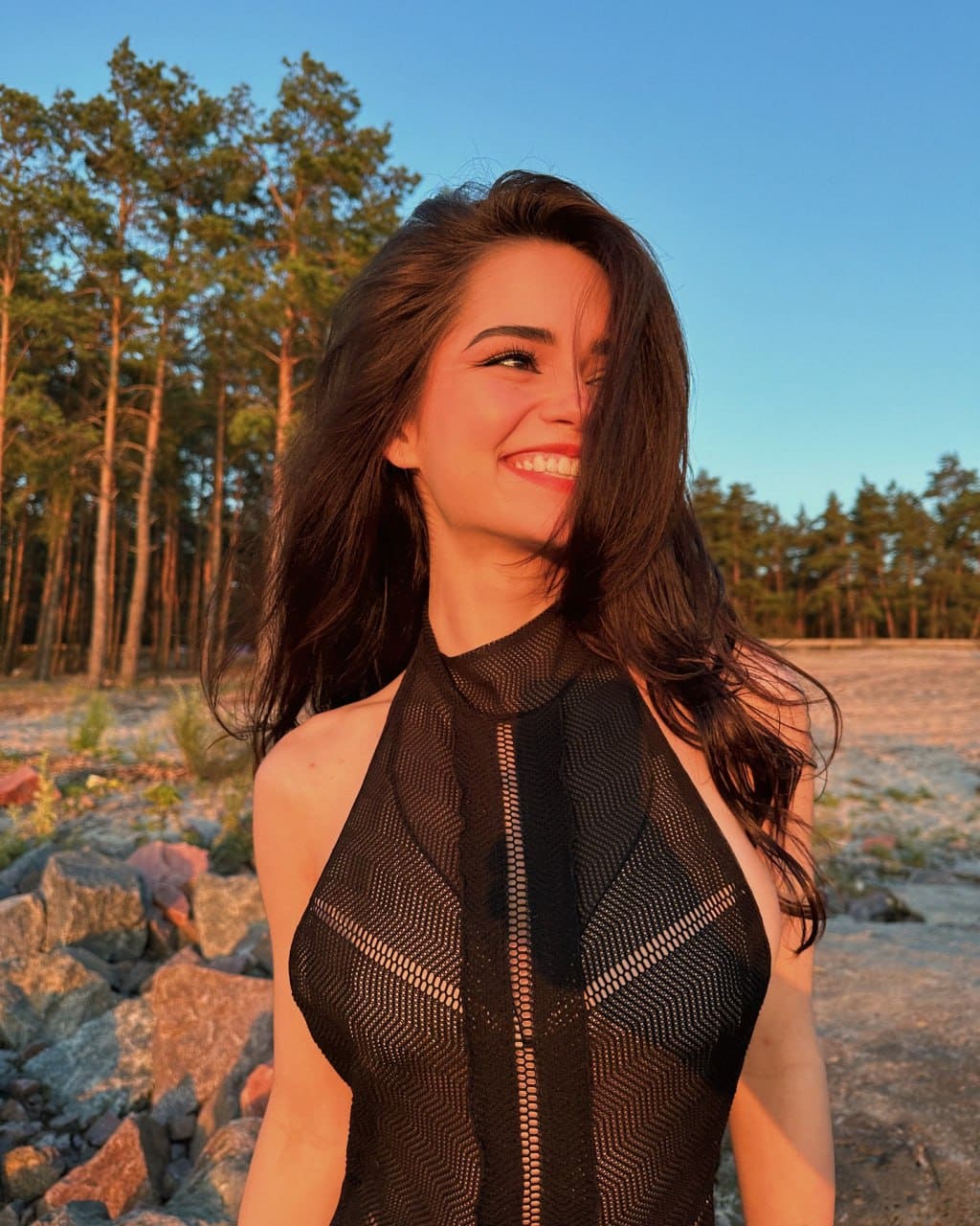 Juliana stands smiling outdoors at dusk before a pine forest, wearing a black halter-neck top with sheer mesh and a central zipper.