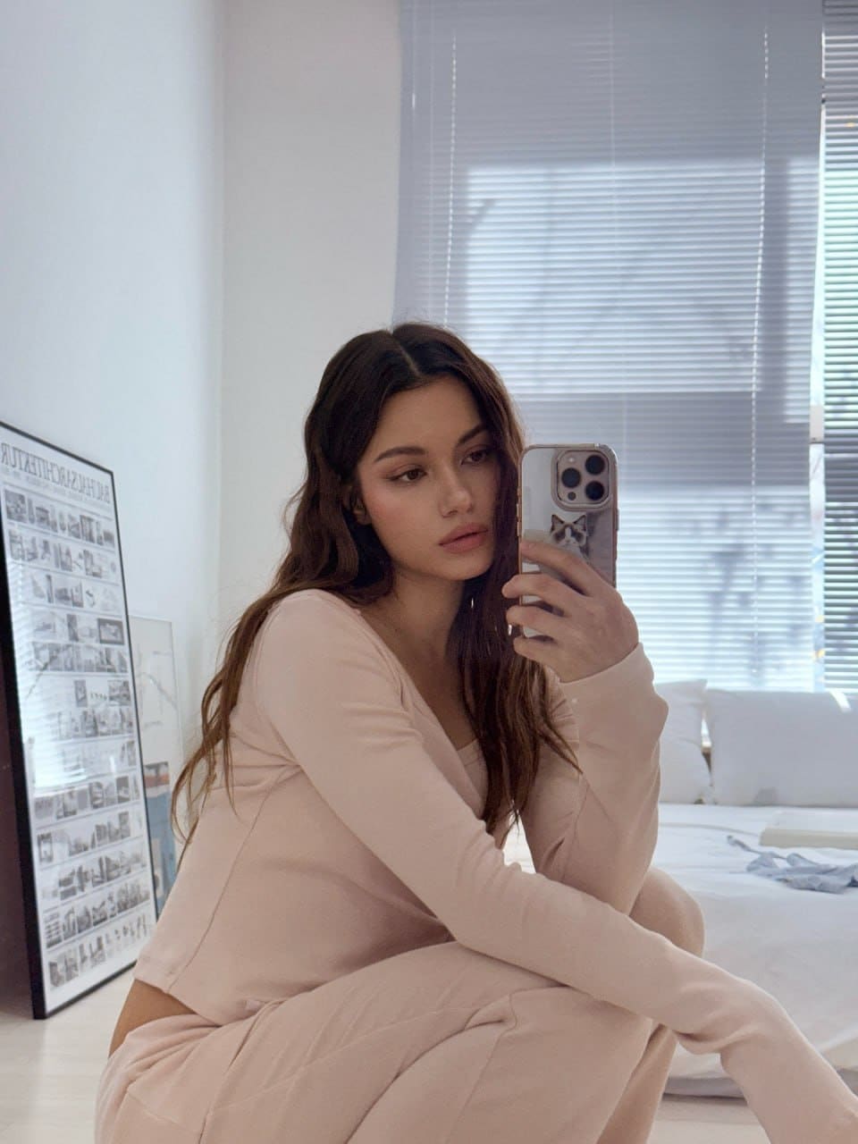 A mirror selfie shows a woman sitting on the floor in a matching light pink long-sleeved top and sweatpants, with a bed and framed poster in the background.