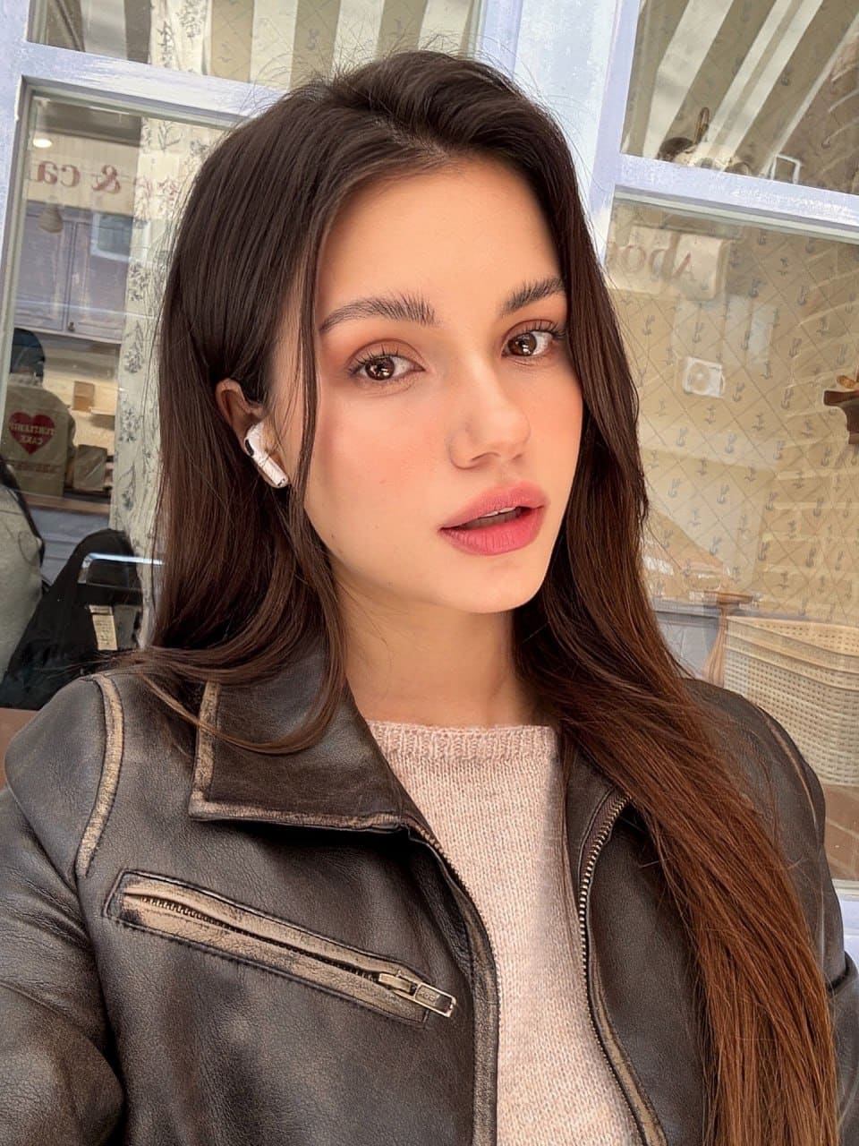 Mira takes a selfie in front of a windowed storefront, wearing a dark leather biker jacket over a light pink knit sweater with a white earbud in her right ear.