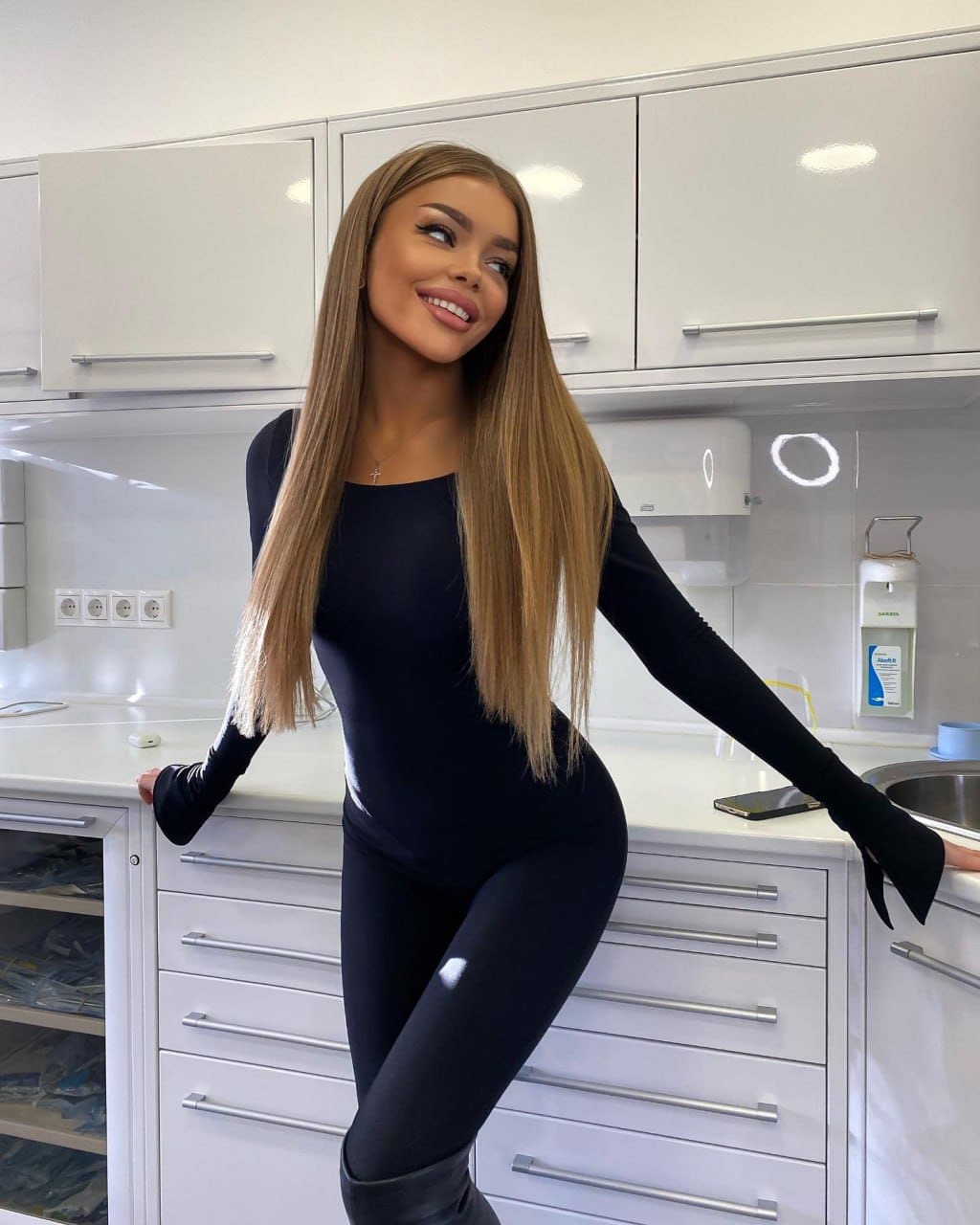 Halia leans against a white kitchen counter in a black bodysuit with thumbholes, black leggings, and tall boots, smiling off to the side.