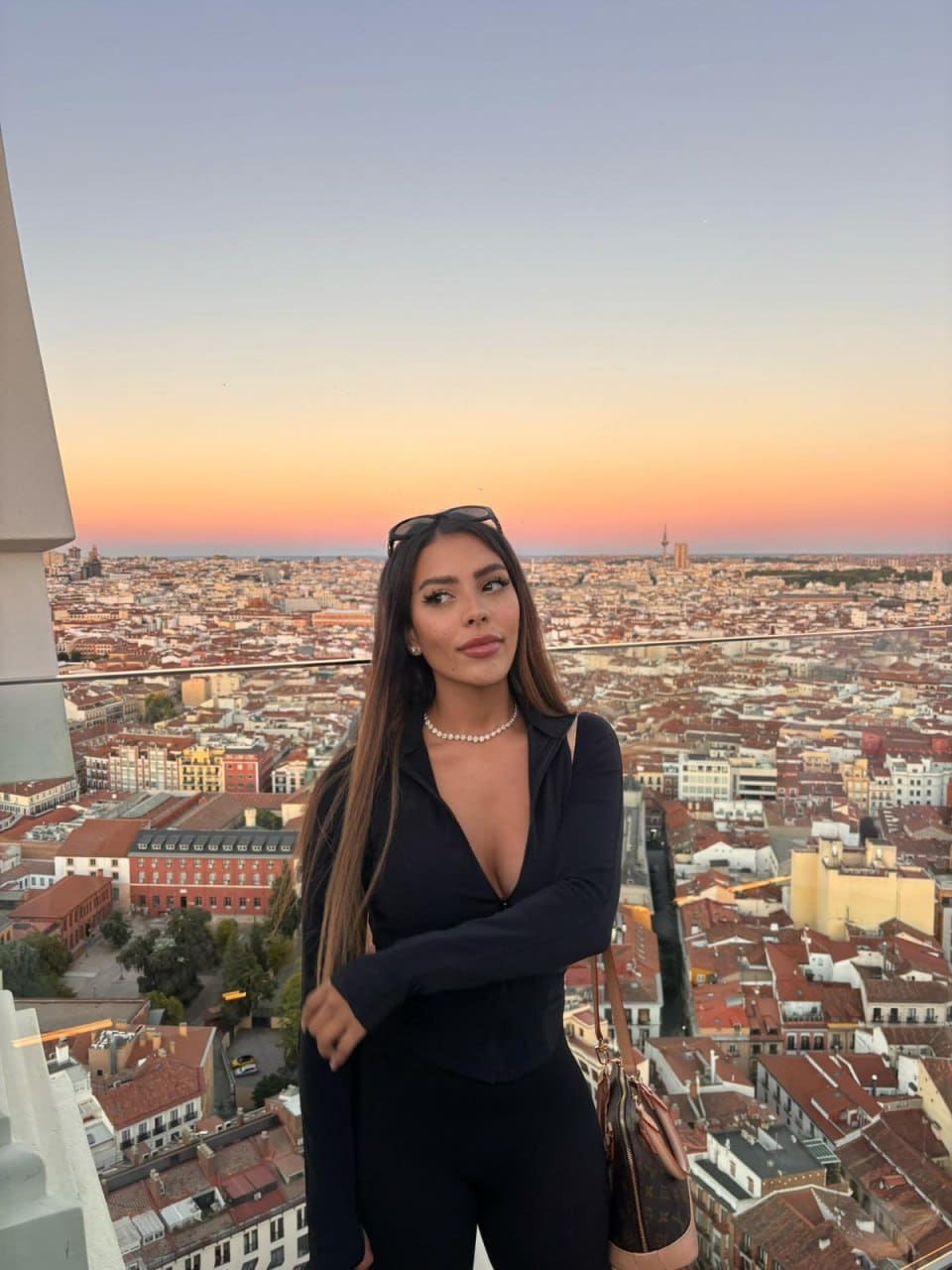 Alana stands on a rooftop terrace at sunset, wearing a black plunging bodysuit, pearl necklace, and sunglasses on her head, holding a handbag.